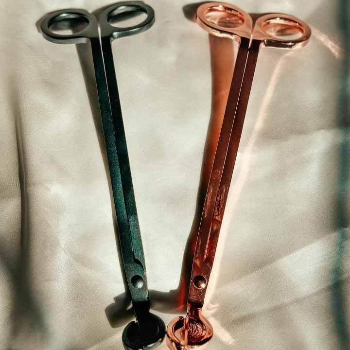 A silver wick trimmer next to a bronze colored wick trimmer. Both lying on a white satin sheet.